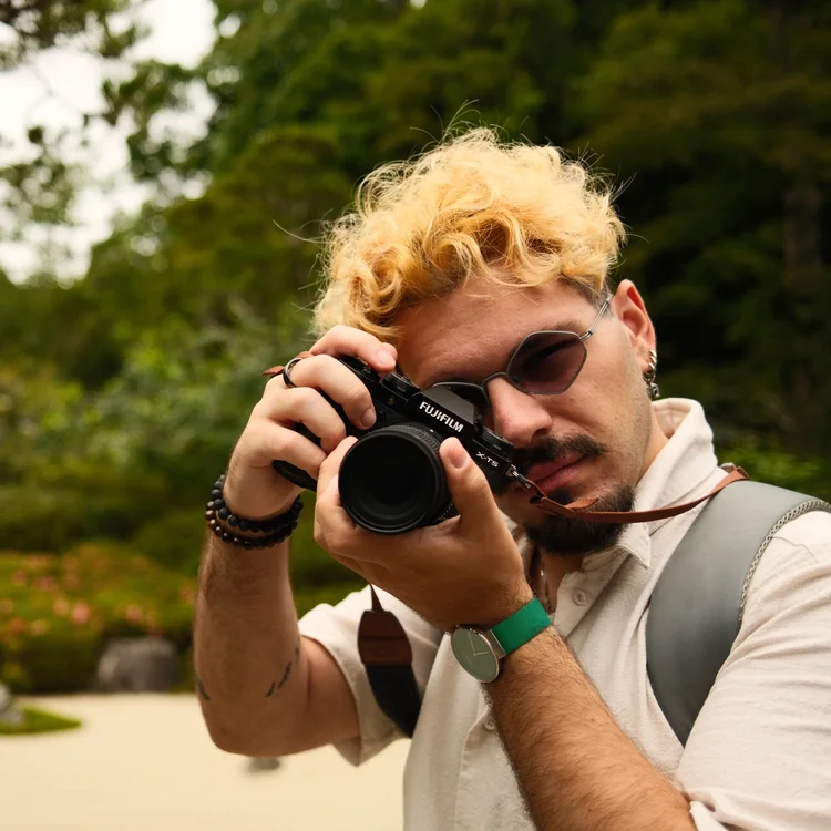 Uomo con capelli ricci biondi, occhiali da sole e t-shirt beige, che scatta una foto con una fotocamera Fujifilm in un ambiente naturale con alberi e acqua sullo sfondo.
